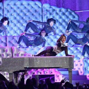 A woman sitting on top of a piano in front of dancers.